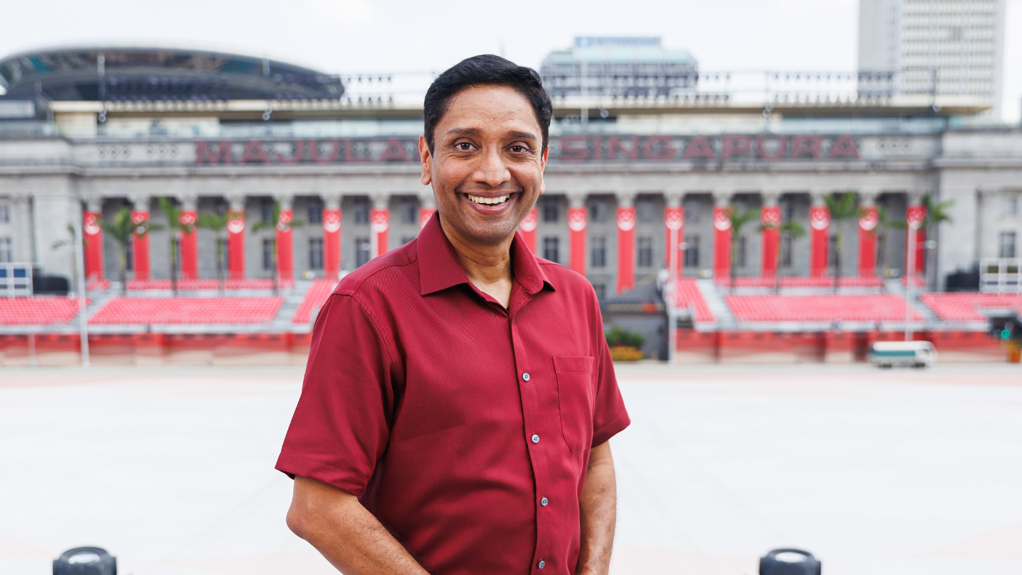 Man in maroon shirt at parade stands; civic building with red national banners behind.